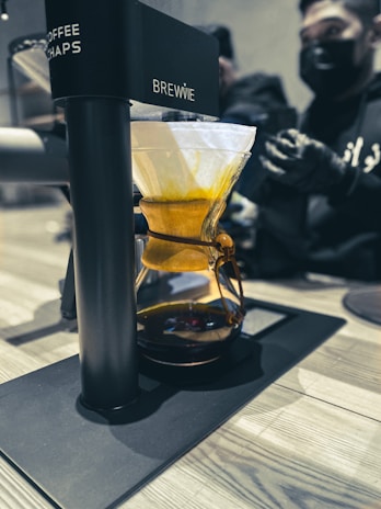 A coffee brewing device labeled 'BREWIE' is situated on a countertop. It features a glass vessel filled with a warm amber liquid and topped with a drip filter holding coffee grounds. In the background, two people wearing black masks and gloves are preparing or observing the process.