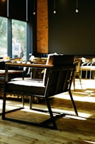 A modern coffee shop interior with wooden floors and dark-colored walls. The space is well-lit with natural light coming through large windows. In the foreground, there is a stylish dark-colored chair with wooden armrests and a sturdy frame. Various other seating options are visible in the background, including chairs and tables. The atmosphere is cozy and inviting, with a blend of modern and rustic elements.