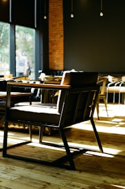 A modern coffee shop interior with wooden floors and dark-colored walls. The space is well-lit with natural light coming through large windows. In the foreground, there is a stylish dark-colored chair with wooden armrests and a sturdy frame. Various other seating options are visible in the background, including chairs and tables. The atmosphere is cozy and inviting, with a blend of modern and rustic elements.