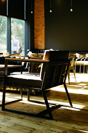 A modern coffee shop interior with wooden floors and dark-colored walls. The space is well-lit with natural light coming through large windows. In the foreground, there is a stylish dark-colored chair with wooden armrests and a sturdy frame. Various other seating options are visible in the background, including chairs and tables. The atmosphere is cozy and inviting, with a blend of modern and rustic elements.