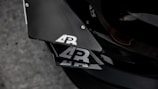 Close-up view of a car's carbon fiber components with a logo that features the letters 'AR' in white on a black background. The surface reflects light, highlighting its sleek and textured appearance. The setting appears industrial with a focus on the angular design and precision of the materials.