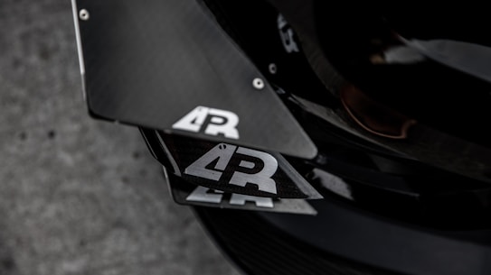 Close-up view of a car's carbon fiber components with a logo that features the letters 'AR' in white on a black background. The surface reflects light, highlighting its sleek and textured appearance. The setting appears industrial with a focus on the angular design and precision of the materials.