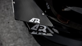 Close-up view of a car's carbon fiber components with a logo that features the letters 'AR' in white on a black background. The surface reflects light, highlighting its sleek and textured appearance. The setting appears industrial with a focus on the angular design and precision of the materials.