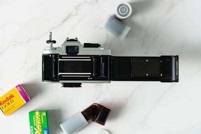An open vintage film camera positioned centrally on a marble surface is surrounded by rolls of film and film canisters. Two film boxes are visible: one with Kodak branding and another labeled Fujifilm.