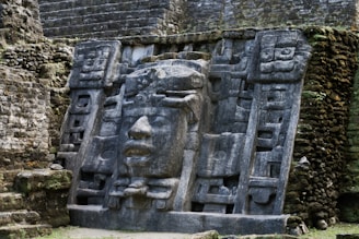 An ancient stone carving depicting a large, expressive face surrounded by intricate geometric designs, set into a weathered stone wall. The sculpture appears to be part of a larger archaeological site, likely of cultural or historical significance.