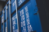 A close-up view of a blue shipping container with white text detailing maximum gross weight, tare weight, and other measurements in both kilograms and pounds. The surface features visible metal reinforcements and a latch system.