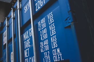 A close-up view of a blue shipping container with white text detailing maximum gross weight, tare weight, and other measurements in both kilograms and pounds. The surface features visible metal reinforcements and a latch system.