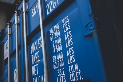 A close-up view of a blue shipping container with white text detailing maximum gross weight, tare weight, and other measurements in both kilograms and pounds. The surface features visible metal reinforcements and a latch system.