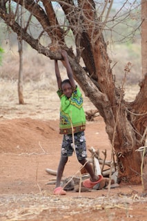 A child wearing a colorful green shirt and patterned shorts stands on a dry, dusty landscape. They are holding onto a branch of a leafless tree, with a background of sparse vegetation and dry soil.