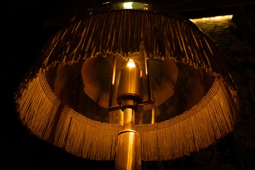 A close-up of a vintage-style lampshade with tassels. Warm light illuminates through the translucent material, casting a cozy glow. The lamp has a metallic base and a single bulb at its center.