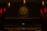 A dimly lit interior scene with cylindrical hanging lights providing soft illumination. A large emblem or logo is mounted prominently on the wall, casting a subtle shadow. Rich red curtains frame the sides, and an illuminated green exit sign is visible below the emblem.