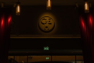 A dimly lit interior scene with cylindrical hanging lights providing soft illumination. A large emblem or logo is mounted prominently on the wall, casting a subtle shadow. Rich red curtains frame the sides, and an illuminated green exit sign is visible below the emblem.