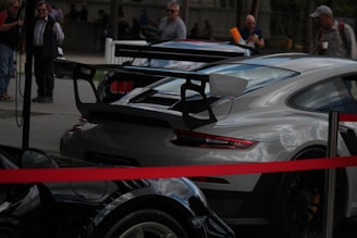 Several high-performance sports cars are parked outdoors, surrounded by a small crowd of people. The cars include models with prominent rear spoilers and sporty designs. A red barrier rope is visible in the foreground.