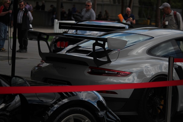 Several high-performance sports cars are parked outdoors, surrounded by a small crowd of people. The cars include models with prominent rear spoilers and sporty designs. A red barrier rope is visible in the foreground.