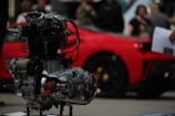A close-up of a mechanical engine component with intricate metal and chrome details. In the blurred background, a vibrant red car is parked, and a few people can be seen, suggesting a car show or exhibition setting.