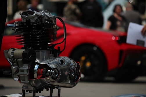 A close-up of a mechanical engine component with intricate metal and chrome details. In the blurred background, a vibrant red car is parked, and a few people can be seen, suggesting a car show or exhibition setting.