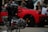 A close-up of a mechanical engine component with intricate metal and chrome details. In the blurred background, a vibrant red car is parked, and a few people can be seen, suggesting a car show or exhibition setting.