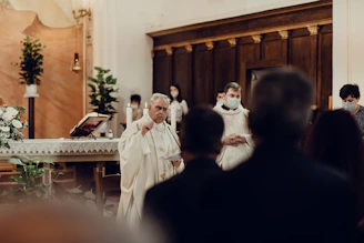 A religious ceremony is taking place inside a church, featuring several people dressed in liturgical vestments. The focus is on an elderly clergy member speaking or reading from a document. Other participants, including some wearing face masks, stand nearby. The altar is adorned with flowers, candles, and an open book, while lush green plants decorate the background.