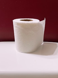 A roll of white toilet paper with an embossed pattern, placed on a smooth white surface, against a deep red wall background.