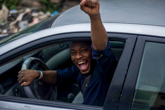 black man in black t-shirt happily waves hand while sitting in driver's seat of car