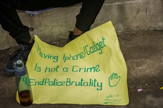 A yellow poster with blue handwritten text is placed on the ground near a person's legs and feet. The text on the poster reads, 'Having iphone/Gadget is not a Crime #EndPoliceBrutality' accompanied by a hand-drawn symbol of an open hand. A plastic water bottle with a dark liquid is sitting nearby on the pavement.