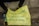 A yellow poster with blue handwritten text is placed on the ground near a person's legs and feet. The text on the poster reads, 'Having iphone/Gadget is not a Crime #EndPoliceBrutality' accompanied by a hand-drawn symbol of an open hand. A plastic water bottle with a dark liquid is sitting nearby on the pavement.