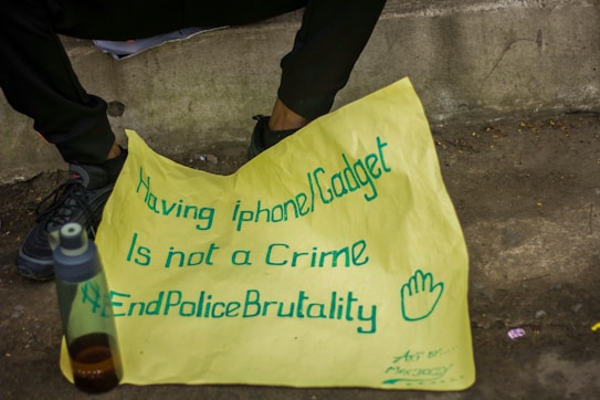 A yellow poster with blue handwritten text is placed on the ground near a person's legs and feet. The text on the poster reads, 'Having iphone/Gadget is not a Crime #EndPoliceBrutality' accompanied by a hand-drawn symbol of an open hand. A plastic water bottle with a dark liquid is sitting nearby on the pavement.