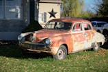 A vintage, rusty car is parked on a lawn in front of a house. The car, showing significant wear and rust, is displayed with a 'For Sale' sign in its window. The house in the background is made of stone and wood, with a large decorative star mounted on the wall. The scene is set in a daytime environment with shadows indicating bright sunlight.