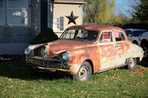 A vintage, rusty car is parked on a lawn in front of a house. The car, showing significant wear and rust, is displayed with a 'For Sale' sign in its window. The house in the background is made of stone and wood, with a large decorative star mounted on the wall. The scene is set in a daytime environment with shadows indicating bright sunlight.