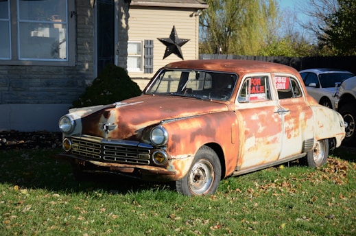 A vintage, rusty car is parked on a lawn in front of a house. The car, showing significant wear and rust, is displayed with a 'For Sale' sign in its window. The house in the background is made of stone and wood, with a large decorative star mounted on the wall. The scene is set in a daytime environment with shadows indicating bright sunlight.