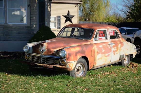 A vintage, rusty car is parked on a lawn in front of a house. The car, showing significant wear and rust, is displayed with a 'For Sale' sign in its window. The house in the background is made of stone and wood, with a large decorative star mounted on the wall. The scene is set in a daytime environment with shadows indicating bright sunlight.