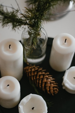 A collection of white pillar candles arranged around a pine cone and a sprig of greenery in a glass jar on a dark surface. The candles are in various stages of being used, with some showing melted wax. The aesthetic appears natural and rustic.