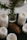 A collection of white pillar candles arranged around a pine cone and a sprig of greenery in a glass jar on a dark surface. The candles are in various stages of being used, with some showing melted wax. The aesthetic appears natural and rustic.