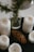 A collection of white pillar candles arranged around a pine cone and a sprig of greenery in a glass jar on a dark surface. The candles are in various stages of being used, with some showing melted wax. The aesthetic appears natural and rustic.