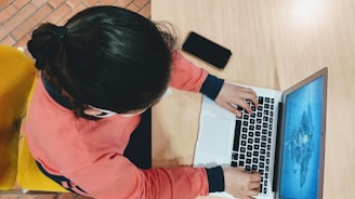 A person wearing a red sweatshirt is typing on a laptop placed on a light wooden table. A smartphone lies nearby, and the person is seated on a yellow chair. The laptop screen displays a blue graphic.