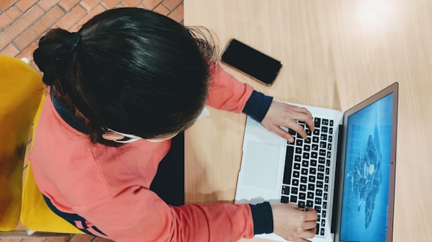 A person wearing a red sweatshirt is typing on a laptop placed on a light wooden table. A smartphone lies nearby, and the person is seated on a yellow chair. The laptop screen displays a blue graphic.