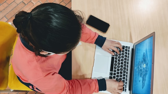 A person wearing a red sweatshirt is typing on a laptop placed on a light wooden table. A smartphone lies nearby, and the person is seated on a yellow chair. The laptop screen displays a blue graphic.