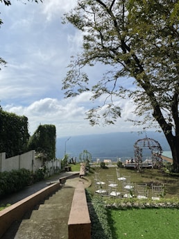 A picturesque outdoor setting featuring a beautiful view of the sky and distant mountains. The scene includes a well-maintained garden with a pathway leading to a terraced area. A large tree with widespread branches adds shade to the setting. Several white chairs are positioned neatly, possibly for an event, along with a decorative arch covered in flowers. The landscape is lush with greenery.