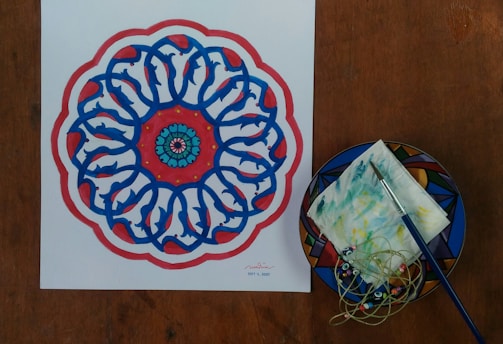 A colorful mandala design featuring intricate blue patterns surrounded by red highlights and a central circular motif with teal and pink accents. Next to the mandala is a plate with a geometric pattern, holding a paintbrush, a napkin smeared with colors, and some tangled cords with beads.