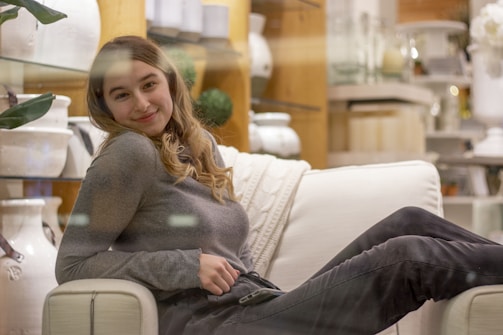 A person with long hair smiling while lounging on a comfortable, light-colored sofa in a store setting. The background features neatly arranged home decor items like vases and potted plants, with wooden shelves providing a cozy, inviting atmosphere.