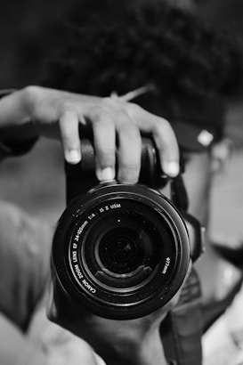 A close-up shot of a person holding a camera with the lens facing forward. The focus is on the camera lens, and the person's hand and part of the face are visible but blurred.