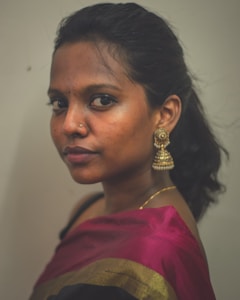 A person is wearing a sari with a border of golden and black colors. The individual has a nose ring and large, ornate earrings. The hair is tied back, and there is a calm expression on the face.
