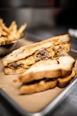 A close-up view of a grilled sandwich filled with melted cheese and cooked ground beef, placed on a tray lined with brown parchment paper. In the background, a bowl of crispy golden fries is slightly blurred.