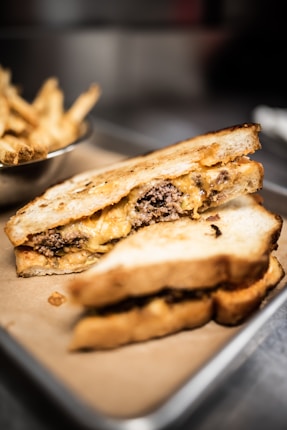 A close-up view of a grilled sandwich filled with melted cheese and cooked ground beef, placed on a tray lined with brown parchment paper. In the background, a bowl of crispy golden fries is slightly blurred.