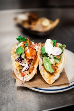 Two delicious tacos filled with ingredients like shredded lettuce, sliced radishes, diced tomatoes, purple cabbage, cilantro, sour cream, and a rich sauce, all served in soft taco shells. They are placed on a piece of brown paper and a white plate with a blue rim. The background is softly blurred, focusing attention on the tacos.
