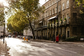 A city street scene with a row of historic brick buildings adorned with greenery and balconies. The street is lined with trees and a few vehicles, including a classic double-decker bus, are on the road. The sunlight casts long shadows and creates a warm ambiance. Pedestrians walk along the sidewalk, and a red postbox stands near the curb.