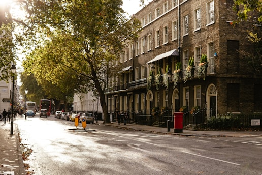 A city street scene with a row of historic brick buildings adorned with greenery and balconies. The street is lined with trees and a few vehicles, including a classic double-decker bus, are on the road. The sunlight casts long shadows and creates a warm ambiance. Pedestrians walk along the sidewalk, and a red postbox stands near the curb.