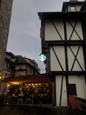 A Tudor-style building with black wooden beams and white walls is prominently featured. Below, a well-lit café with people seated at tables under a maroon canopy is visible. The café has a green and white coffeehouse logo, hanging from the building. The sky is overcast, giving the scene a moody atmosphere.
