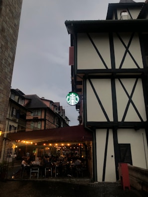 A Tudor-style building with black wooden beams and white walls is prominently featured. Below, a well-lit café with people seated at tables under a maroon canopy is visible. The café has a green and white coffeehouse logo, hanging from the building. The sky is overcast, giving the scene a moody atmosphere.