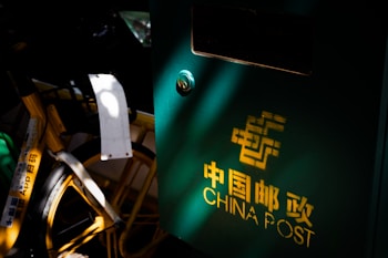 A green China Post mailbox is prominently shown, with light casting shadows and highlighting a portion of the logo in bright yellow. Adjacent to it, the wheel and part of a yellow bicycle are visible, with some Chinese characters and a white board attached to the bike, partially illuminated.